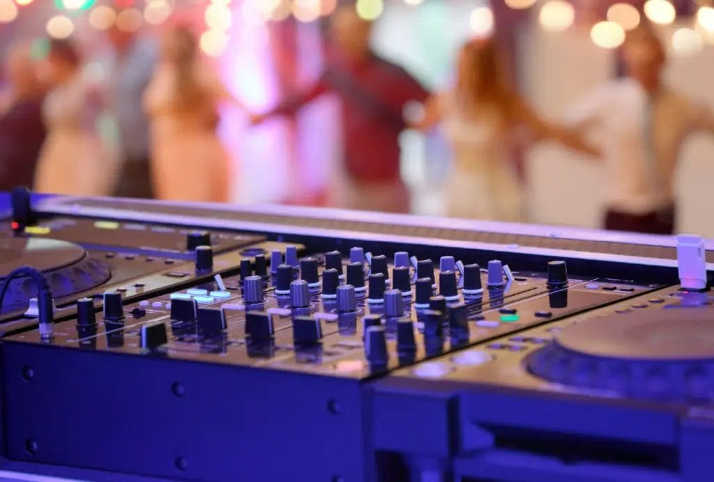 A DJ mixer with wedding guests dancing in the background.
