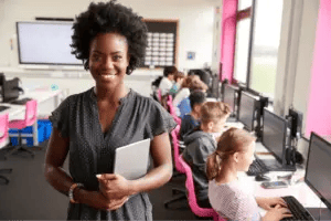 Teacher holding a tablet stands in front of her class, who is working on computers in a school computer lab