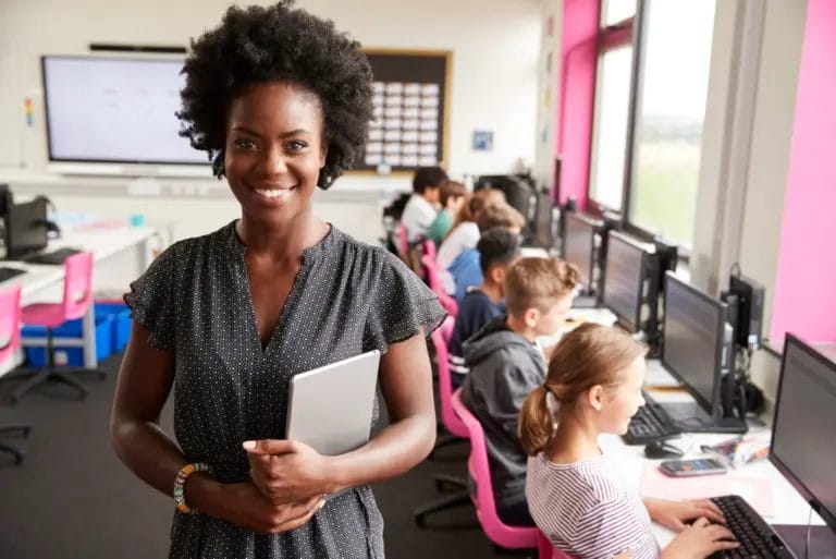 Teacher holding a tablet stands in front of her class, who is working on computers in a school computer lab