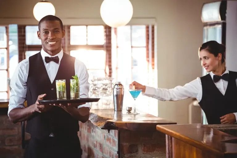 A male bartender holds a tray with cocktails and smiles at the camera while a female bartender finishes making a cocktail at an indoor bar during an event.