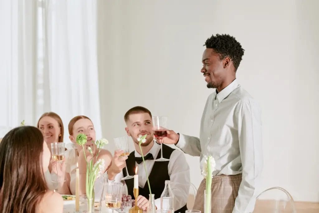 a group of men and women sitting at a wedding celebration table smiling and raising glasses while a man stands and gives a toast