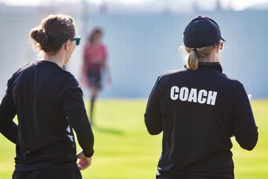 Rear view of two coaches watching players on a field.