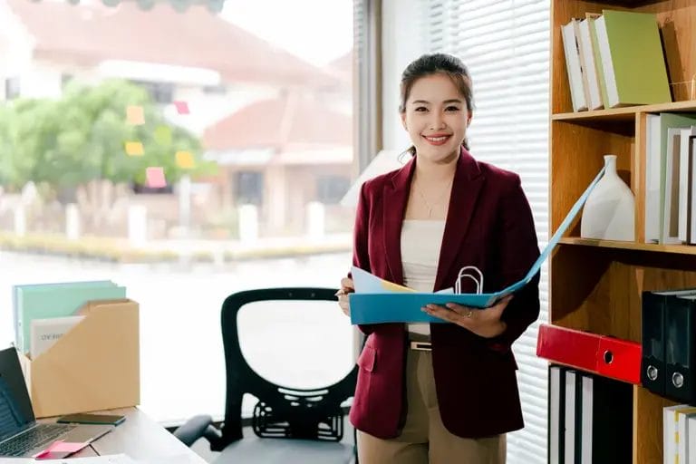 A consultant in a burgundy blazer holds a blue binder and smiles at the camera in a school office.