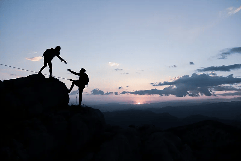 Silhouetted against a sunrise, one rock climber extends a hand to help another summit a peak.