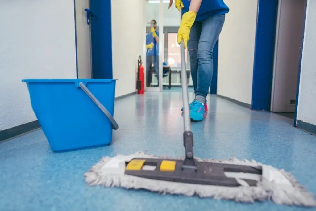 a close-up of a cleaning employee mopping a hallway floor while another employee cleans windows