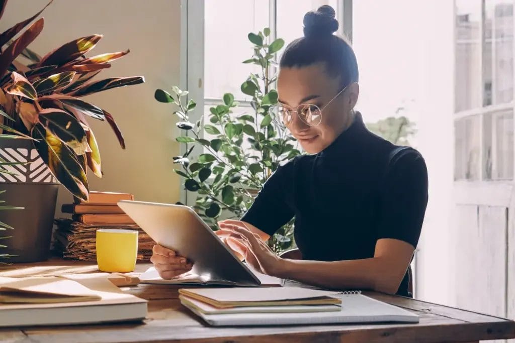 A small business owner wearing glasses and a black turtleneck blouse taps on a business laptop at a desk decorated with houseplants near an open door with sunlight spilling in.