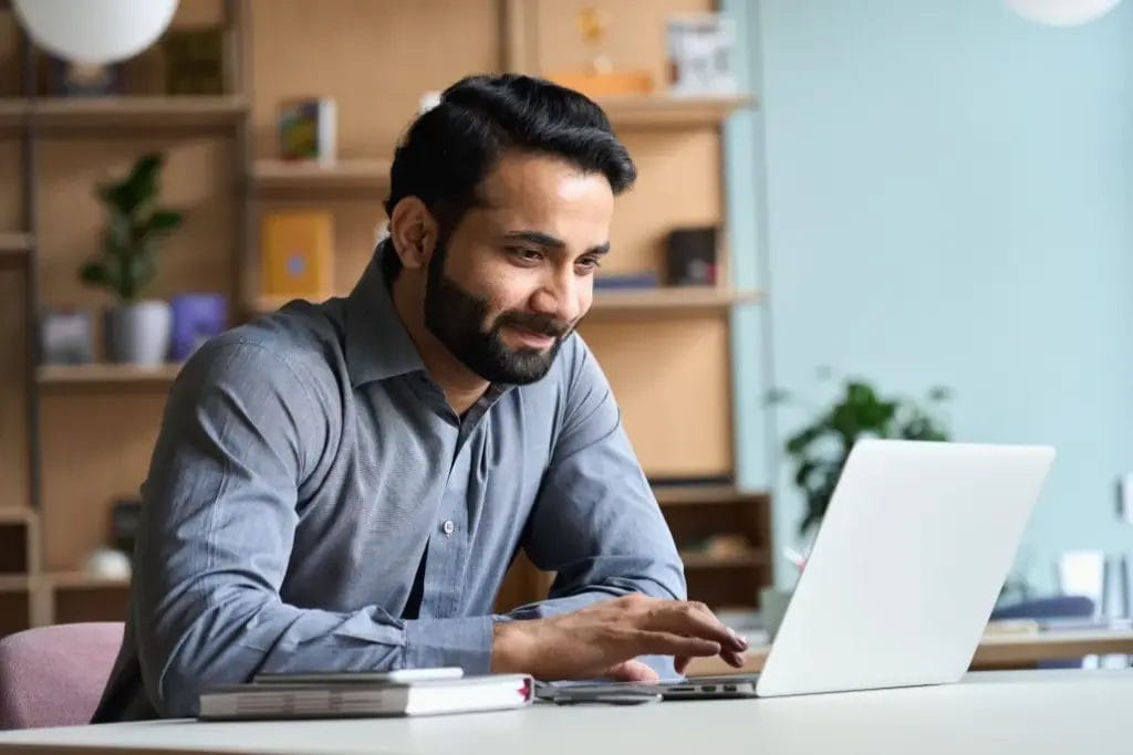 A small business owner wearing a gray button-up shirt types on his business laptop in a room with a large wooden shelving unit behind him, decorated with plants and artwork.