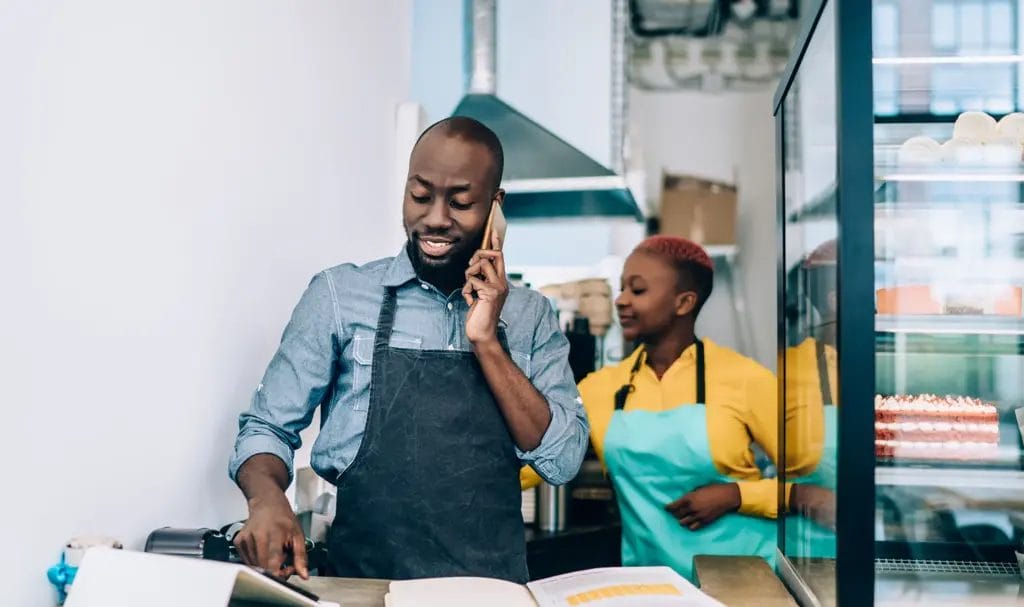 A baker is taking an order from a customer on the phone while an employee helps prep ingredients in the background.