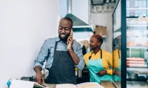 A baker is taking an order from a customer on the phone while an employee helps prep ingredients in the background.