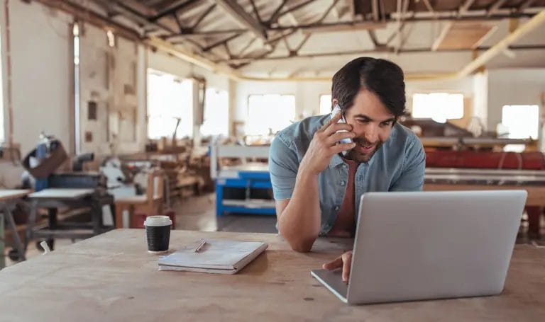 Small business owner talking on the phone using a laptop at their workbench in a small manufacturing studio space.