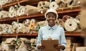 A small business owner smiles while wearing a white hard hat and holding a clipboard as she stands by shelves holding large fabric rolls.