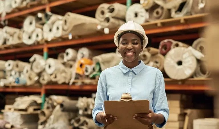 A small business owner smiles while wearing a white hard hat and holding a clipboard as she stands by shelves holding large fabric rolls.