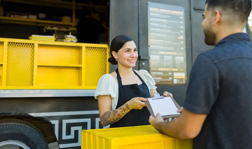 A food truck owner happily shares paperwork on a clipboard with another person.