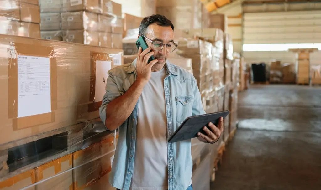 A small business owner talks on the phone while reading information on a tablet and standing next to large shipment boxes in a warehouse.