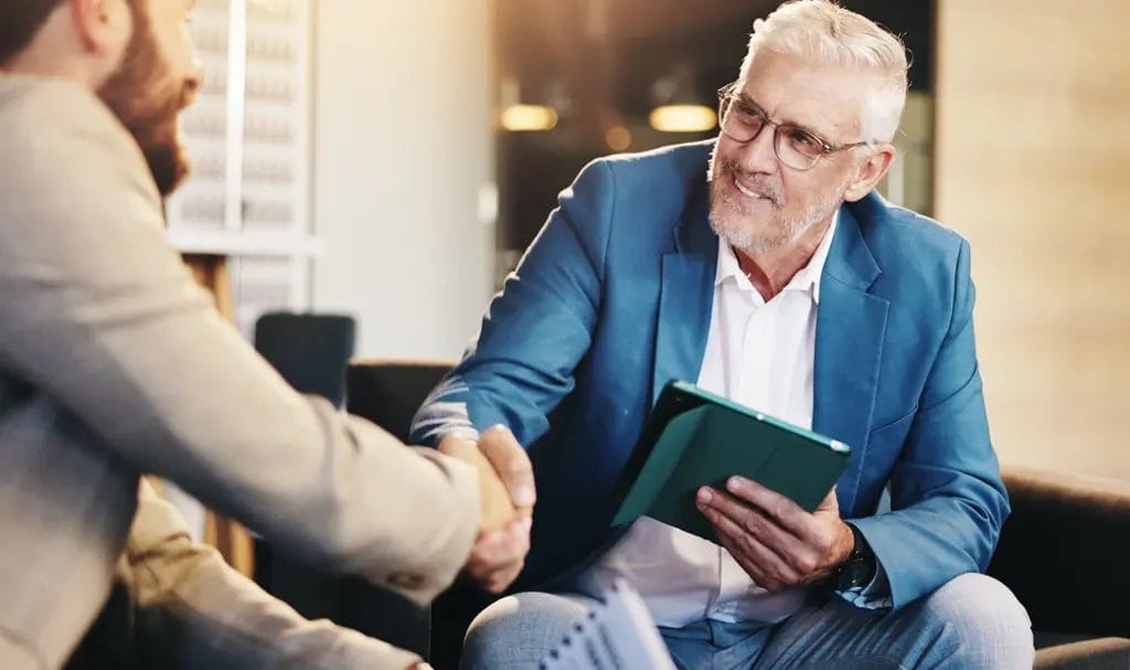 A businessman happily shakes hands with a new business partner with one hand as he holds an ipad in his other hand after reviewing contract details.