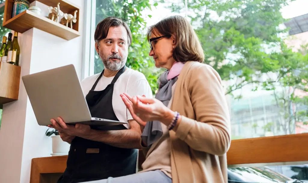 A small business owner and customer having a discussion about while the business owner holds a laptop between the two of them.