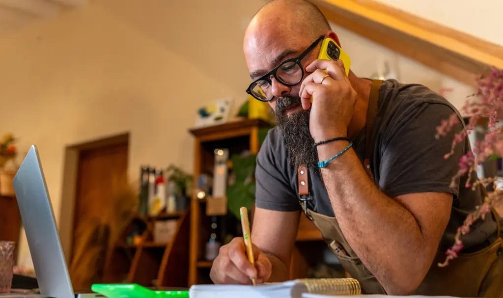 Business owner in his workspace speaking on the phone while taking notes in a notebook on a desk next to an open laptop.