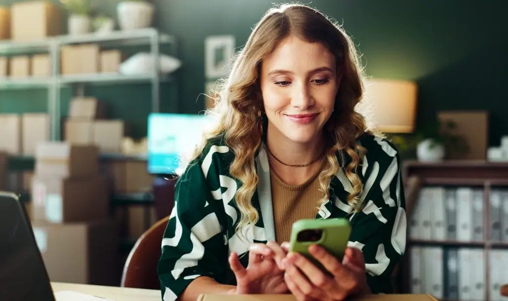 Small business owner smiling while using a smartphone in their home office.