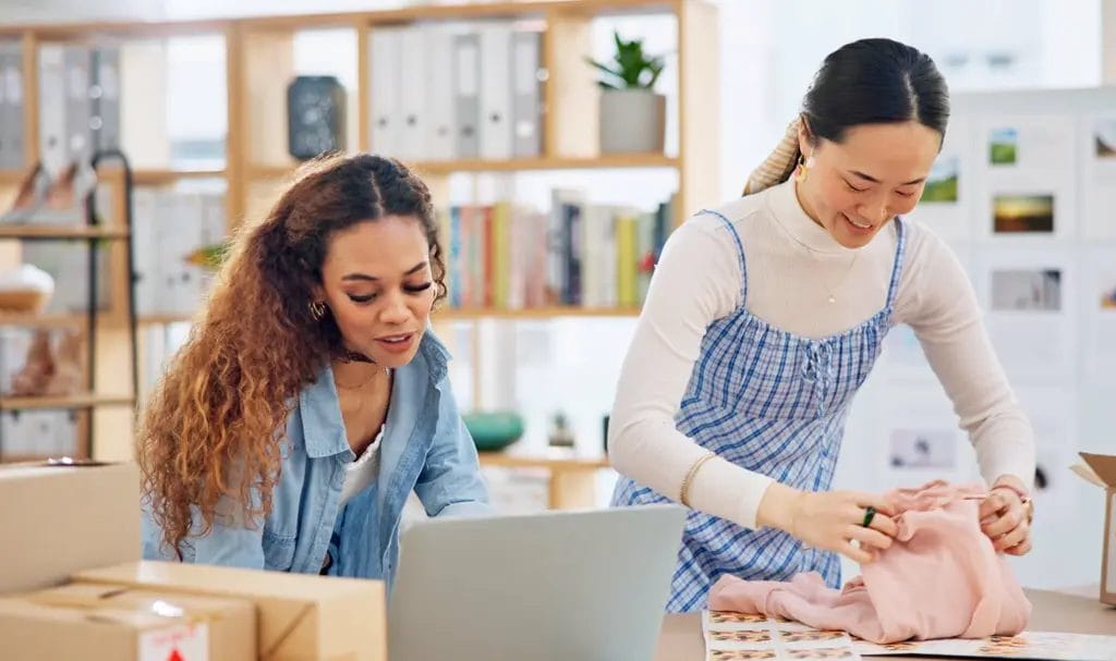 Two businesswomen are discussing contract details on a laptop as one sits next to packaged items ready to be shipped and the other is folding clothing items ready to be packaged.