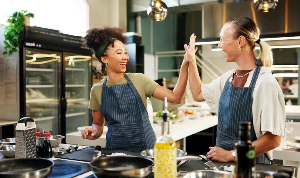 A chef happily high-fives her employee as they begin prepping a catering order in a commissary kitchen.