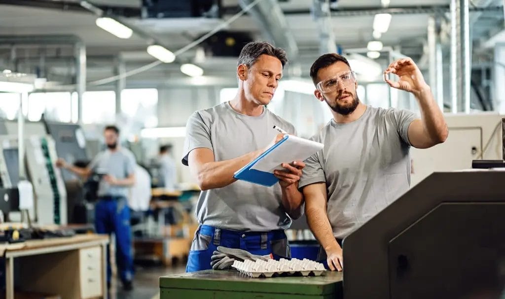 Two employees are inspecting parts being manufactured in a warehouse with one taking notes on a clipboard.