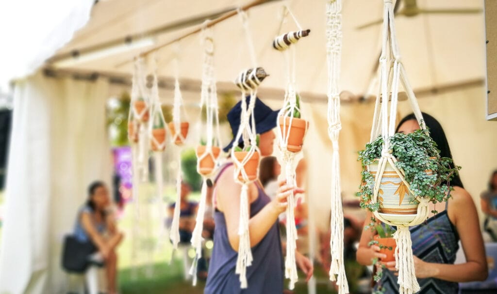 Buyers look at plants hanging in handmade crocheted baskets hanging from a vendor's booth tent.