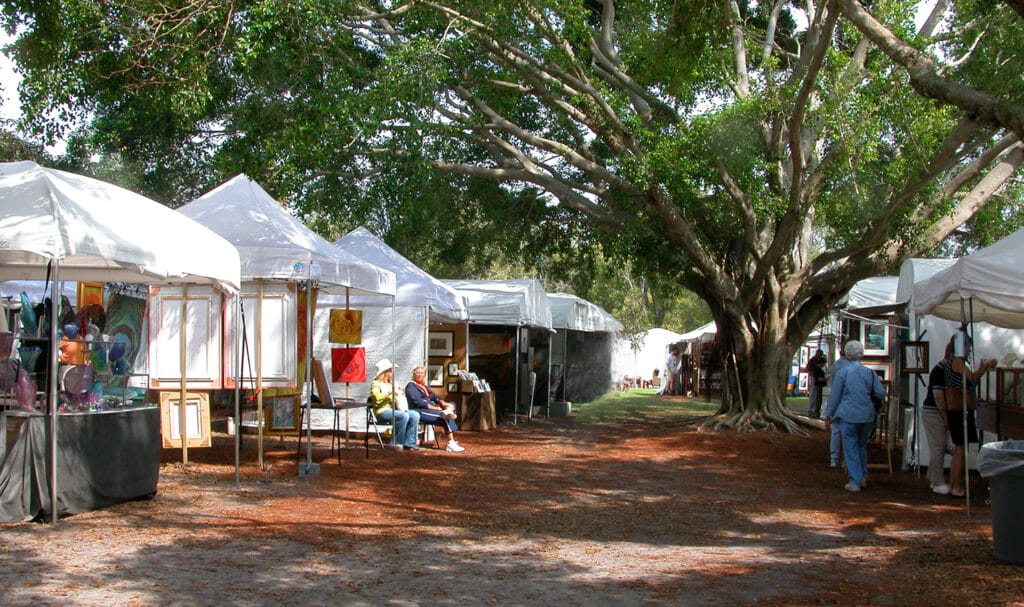 Artists and crafts displaying their work in vendor tents at an outdoor event in a shady park.