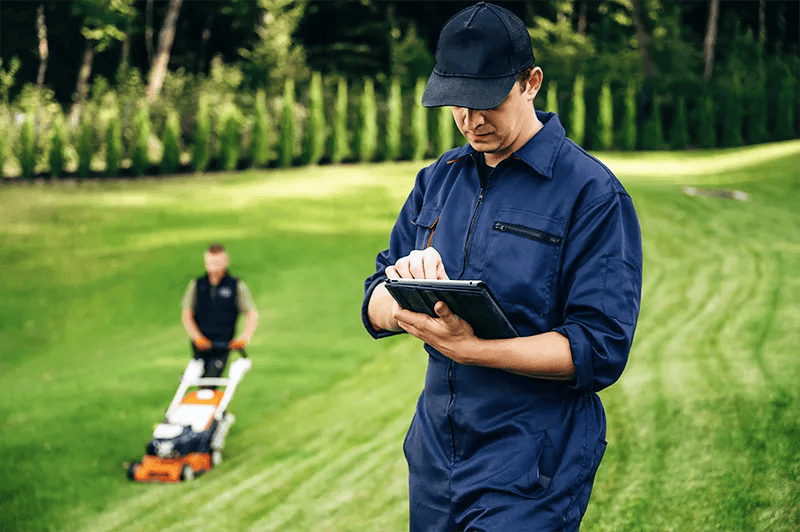 Image of a lawn care professional using an IPad with his partner mowing the lawn in the background.