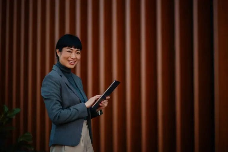 A small business owner wearing a teal turtleneck and blazer smiles while holding a laptop and walking in front of a red slatted building.