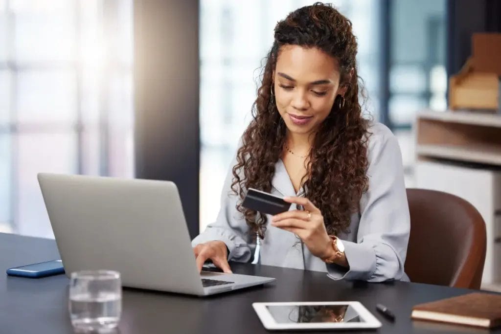 A small business owner wearing a gray blouse looks at her credit card details while typing at her business laptop as she sets up an account.