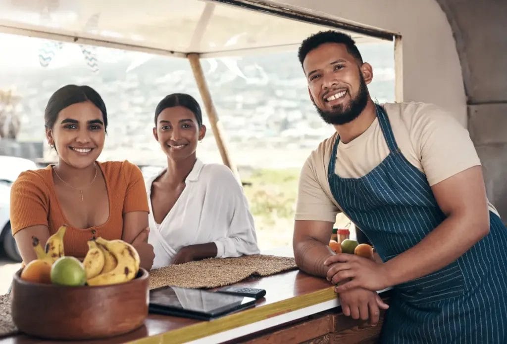 A food truck owner wearing a denim striped apron smiles and leans into a food truck window with two workers; the window is decorated with rattan placemats and a bowl of fruit.