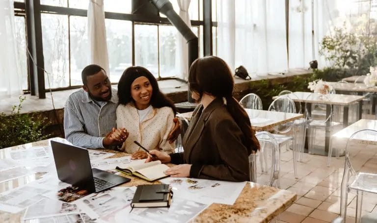 A couple seated at a table holds hands and excitedly listens to a venue manager walk them through details of a wedding venue contract.