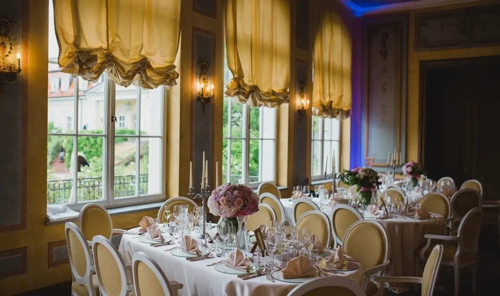 A room in a historic building has tables and chairs set for a wedding reception, with floral arrangements and candelabras adorning each tabletop.