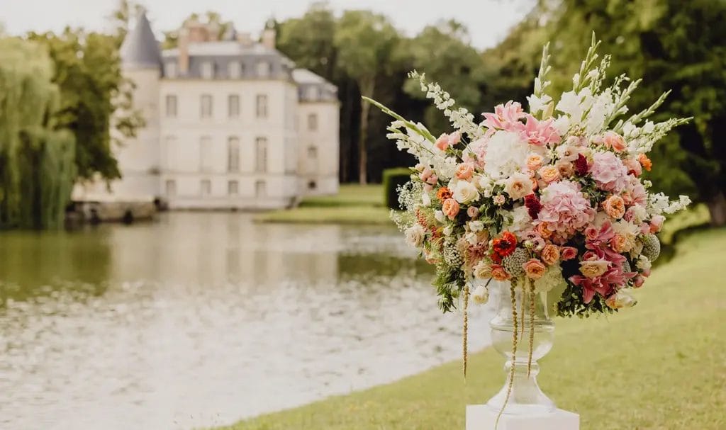 A wedding floral arrangement sits on a white pillar, with a private estate and lake in the background.