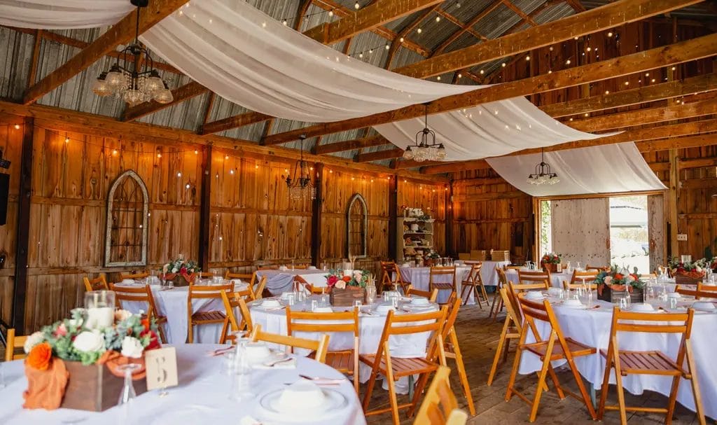 A rustic barn serves as a wedding venue, with reception tables, string lights, and chandeliers ready for the occasion.