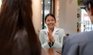 A young consultant happily looks across a table at two of her clients as she smiles and clasps her hands in excitement.