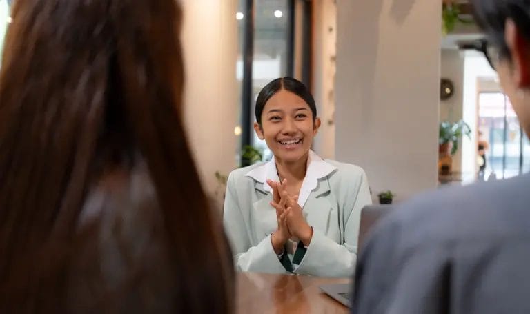 A young consultant happily looks across a table at two of her clients as she smiles and clasps her hands in excitement.