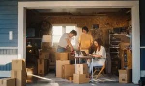 A group of small business partners works from their set-up in a garage. One is typing on a laptop at a small desk, while the other two are packaging products for shipment.