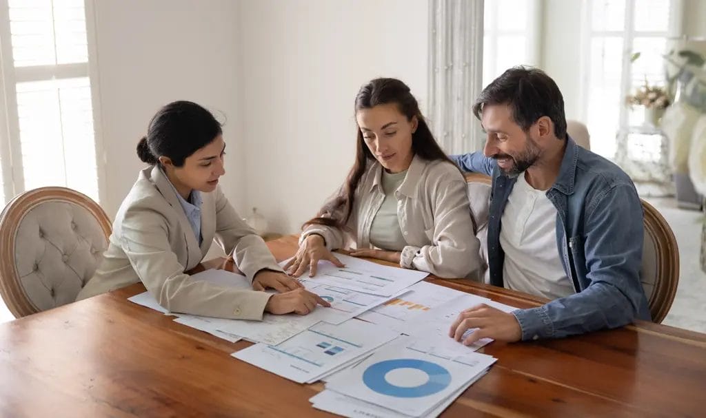 An advisor walks two clients through paperwork and data information at a large home dining table.