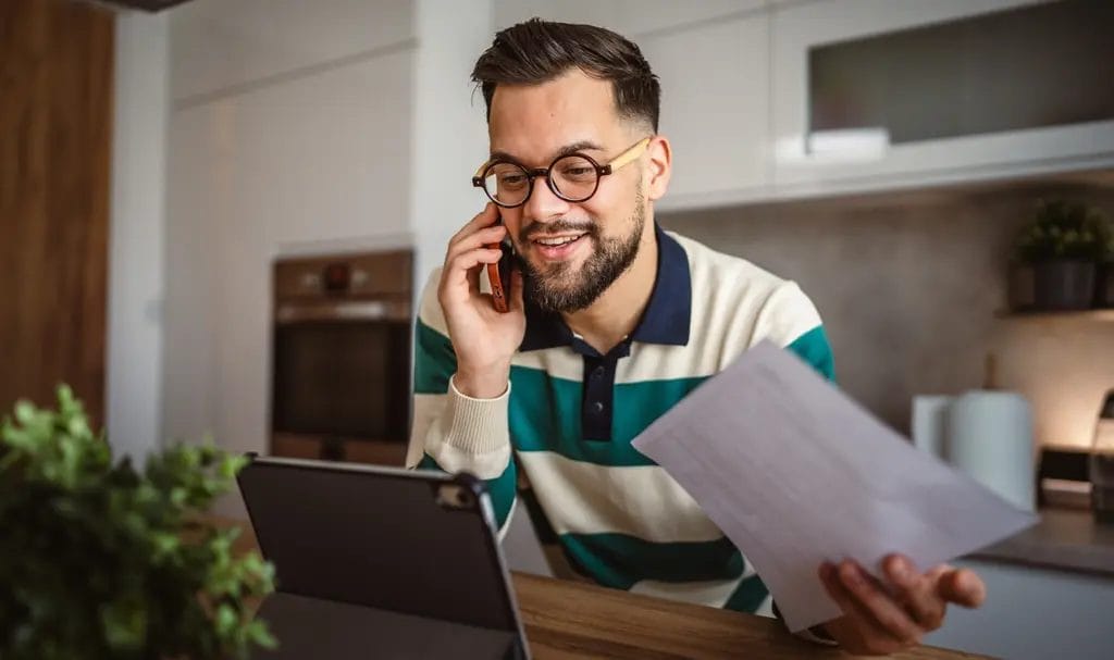 A small business owner cheerfully speaks on the phone while looking at a tablet propped up on his counter and holding paperwork in his other hand.