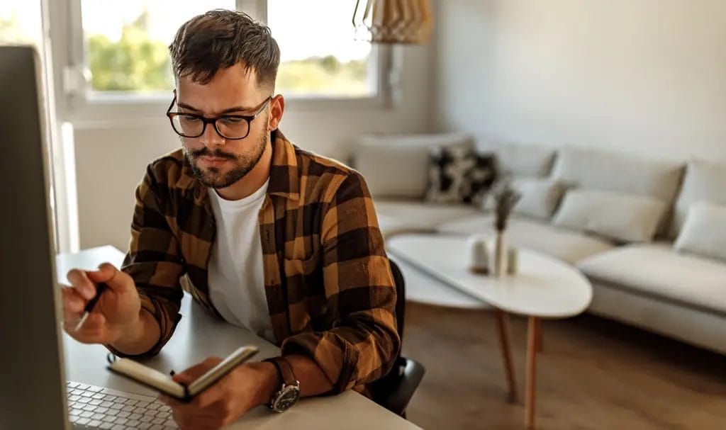 A professional designer reviews notes in a notebook after a call with a client as he sits in front of a computer in his home.