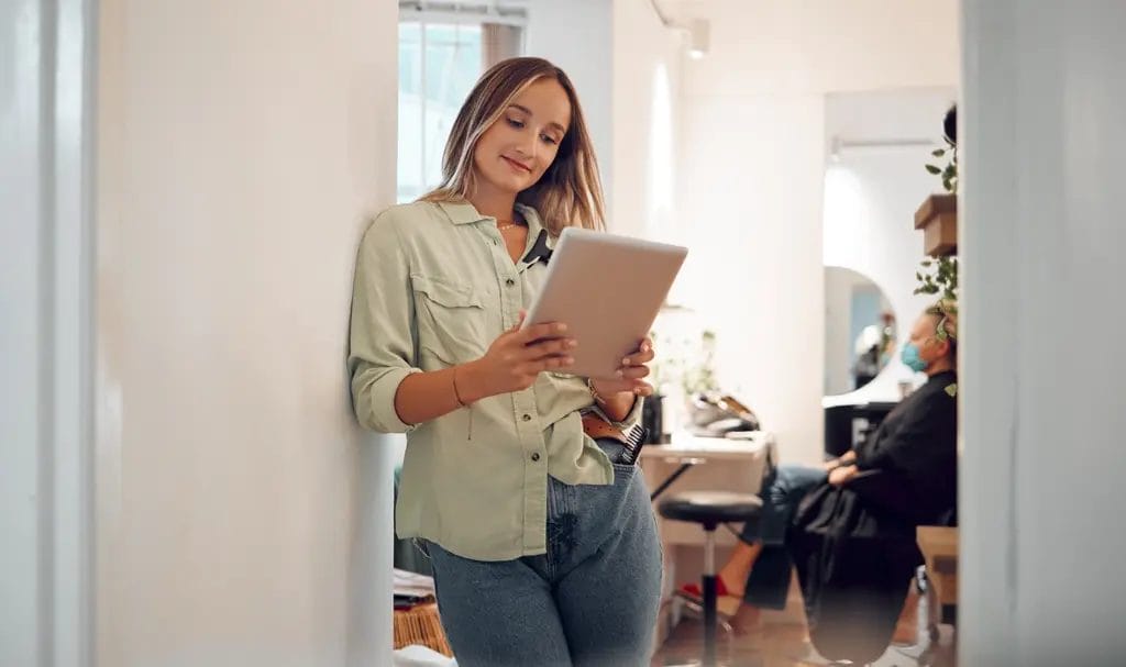 A hairstylist leans against a wall while she reviews business contracts on an tablet.