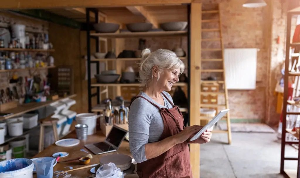 A small business crafter smiles as she leans against her workbench in her craft studio and reviews information on a tablet.