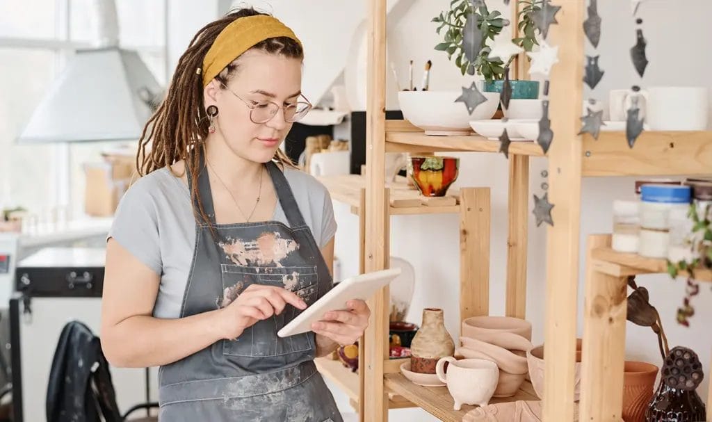 A small business crafter is reviewing order details on a tablet in her pottery studio next to a shelf of various handmade clay wares.