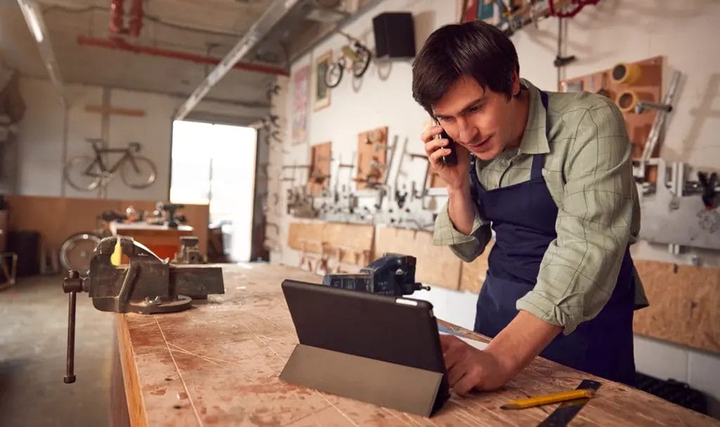 A small business repairman is speaking on the phone while scrolling through information on a tablet that is propped up on his workbench.