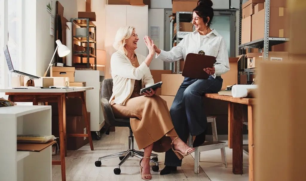 A pair of businesswomen high-fives while sitting in a small workspace surrounded by shipping boxes on shelves.