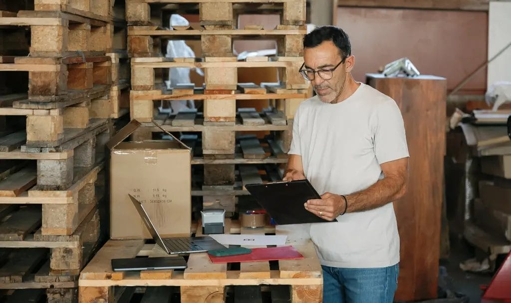 A businessman reviews a contract next to his laptop and other paperwork that he has set on a makeshift desk of stacked wooden pallets in a warehouse.