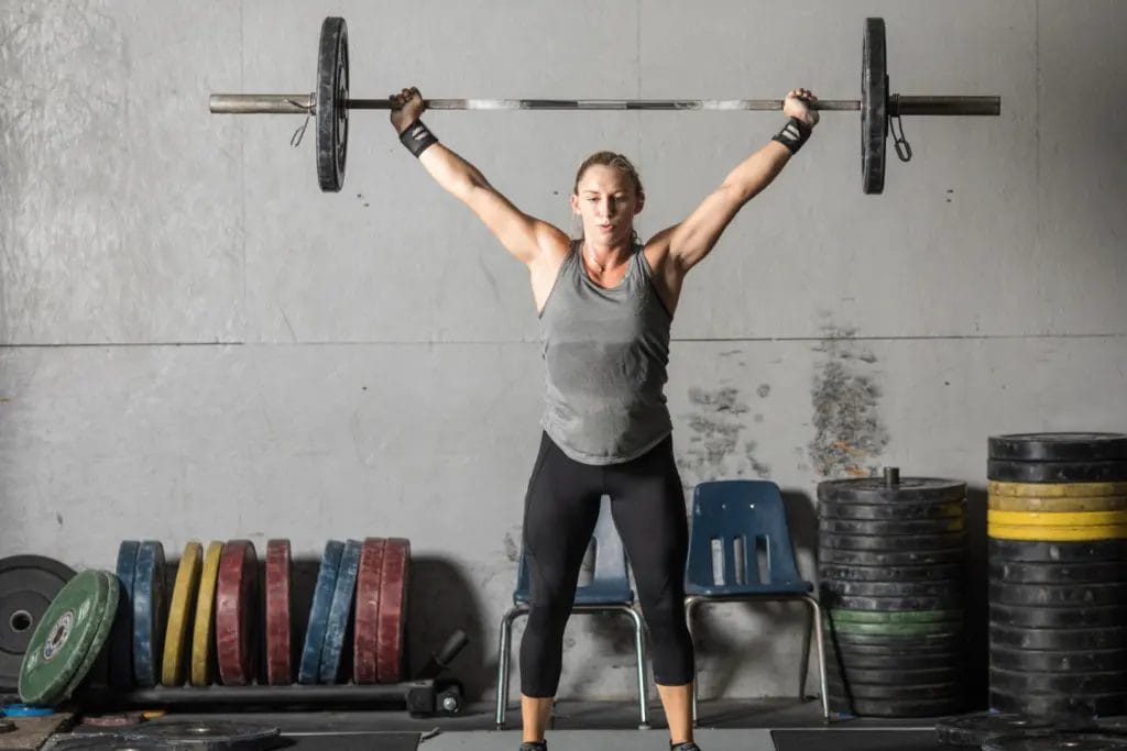 A woman performing a barbell snatch, holding the barbell overhead inside a weightlifting gym.