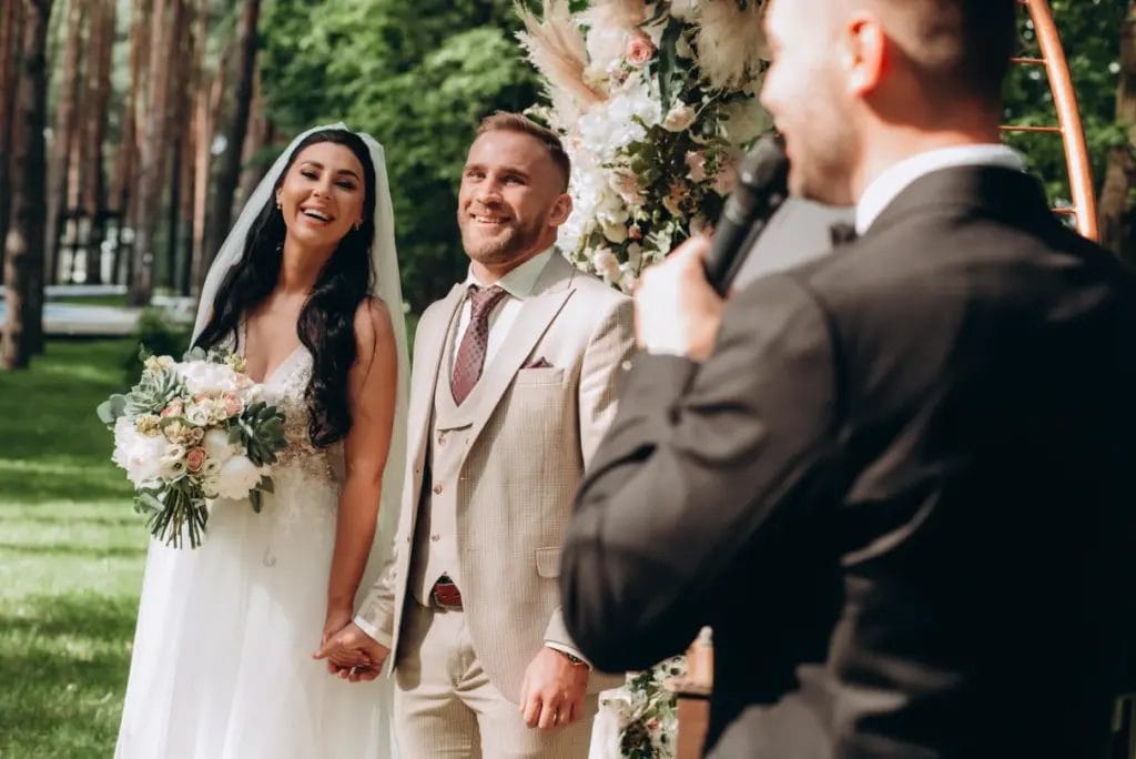 a wedding host speaks into a microphone while addressing a bride and groom in front of a floral arch