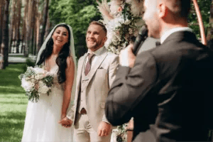 a wedding host speaks into a microphone while addressing a bride and groom in front of a floral arch